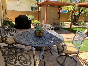 Een terras met een ronde metalen tafel en stoelen, omgeven door potplanten en een barbecue.