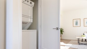 A room with a white door, a washing machine, a small table with a plant, and two framed pictures on the wall.