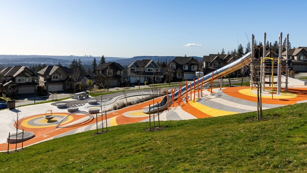 A playground with a slide and a curved path, surrounded by houses and a clear sky.