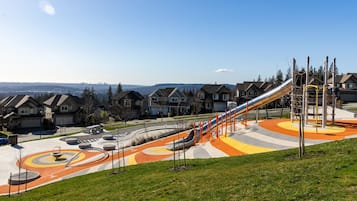 A playground with a slide and a curved path, surrounded by houses and a clear sky.