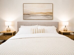 Un lit bien fait, avec une couette à motifs, deux lampes de chevet et un tableau abstrait encadré au-dessus du lit.