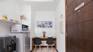 Una kitchenette compacta con microondas, refrigerador y dispensador de agua. Hay una pequeña mesa de comedor con dos sillas y un florero con plantas secas. Se ve un cartel de 'No Smoking' en la pared.