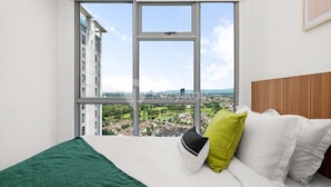 Quarto com uma grande janela, cama com roupa de cama branca e verde, e vista para a paisagem urbana.
