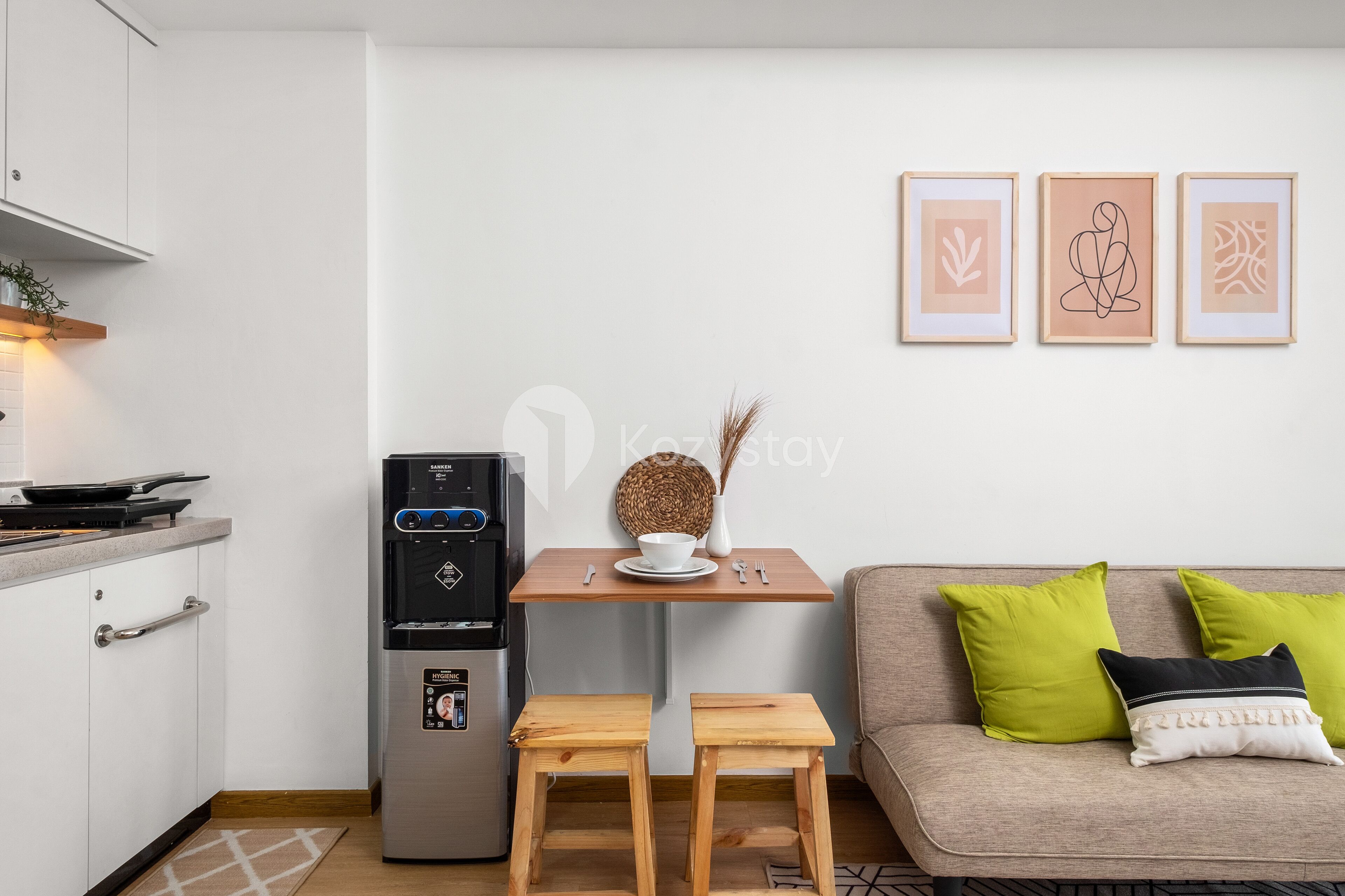 A compact kitchen with a refrigerator, a small table with a bowl and a vase, and a sofa with green cushions.
