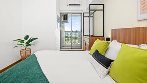 Quarto de hotel com cama, planta em vaso, varanda e vista, e cabeceira de madeira.
