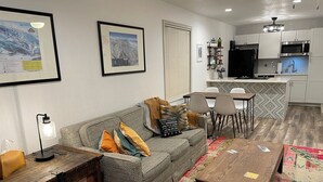 Uma sala de estar moderna com sofá, mesa de centro e área de jantar. Há uma cozinha com armários e eletrodomésticos brancos.