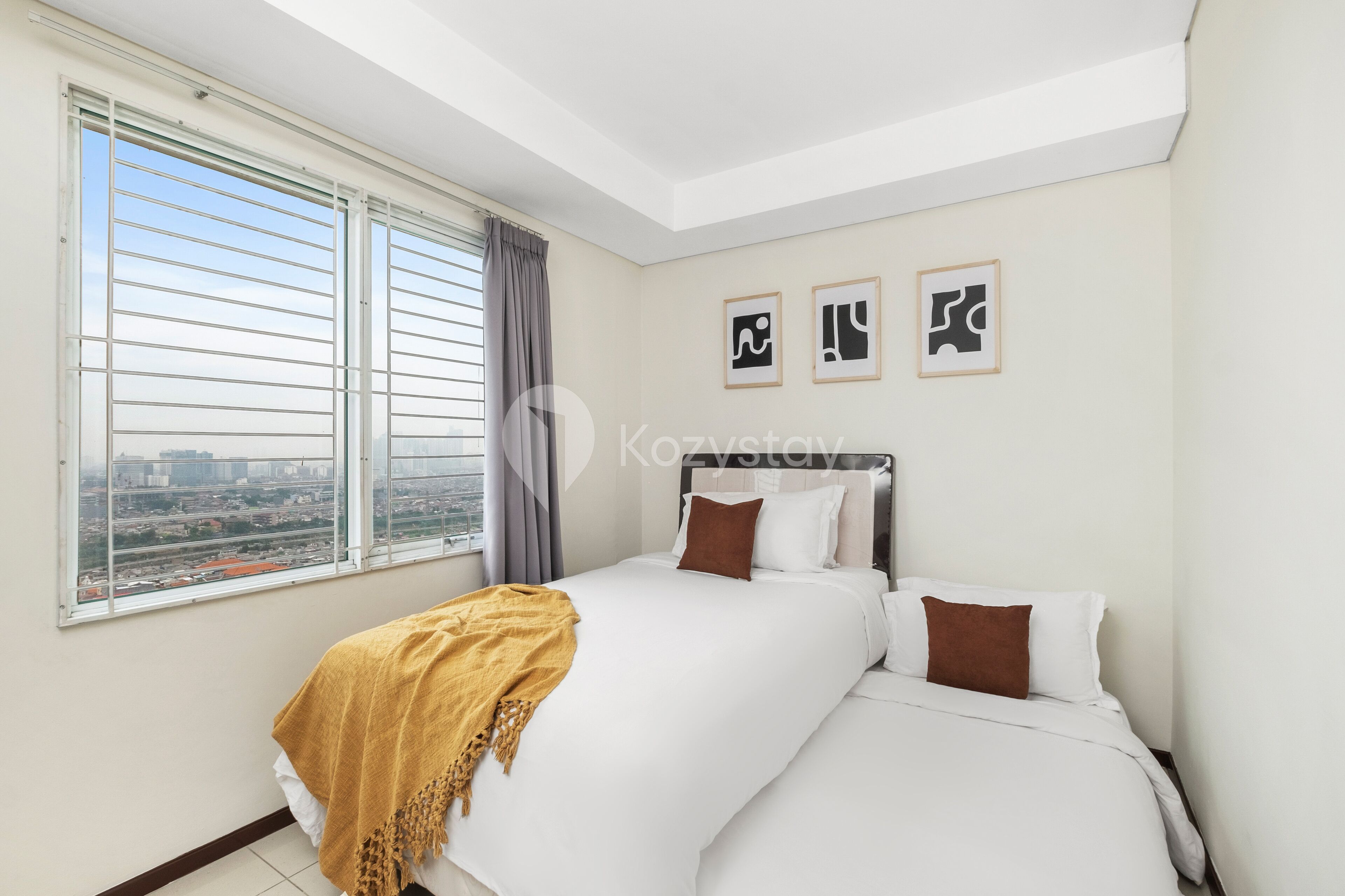 A bedroom with a large bed, a view of the city through a window, and three framed pictures on the wall.