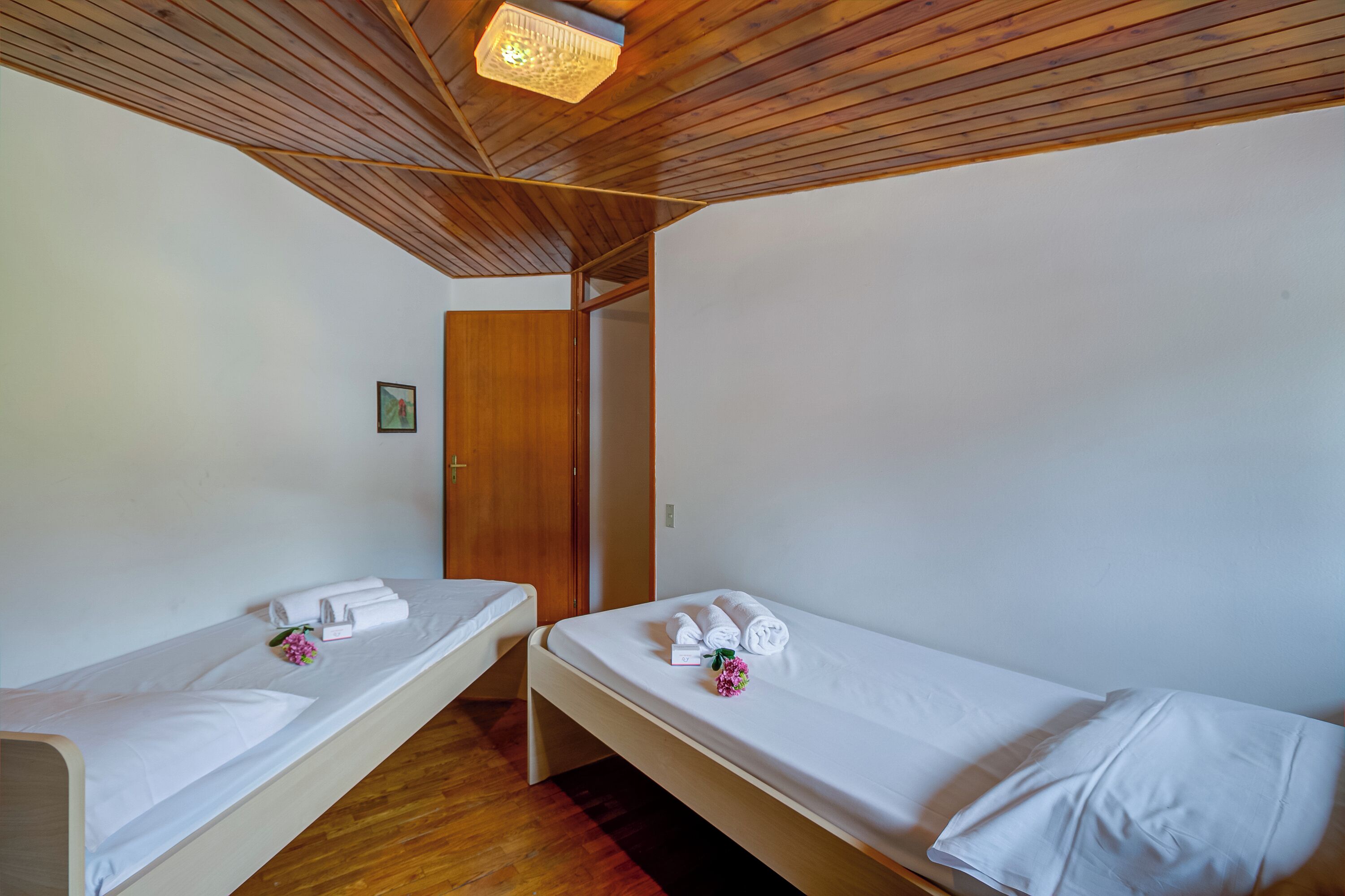 A room with two single beds, wooden ceiling, white walls, and a small framed picture on the wall.