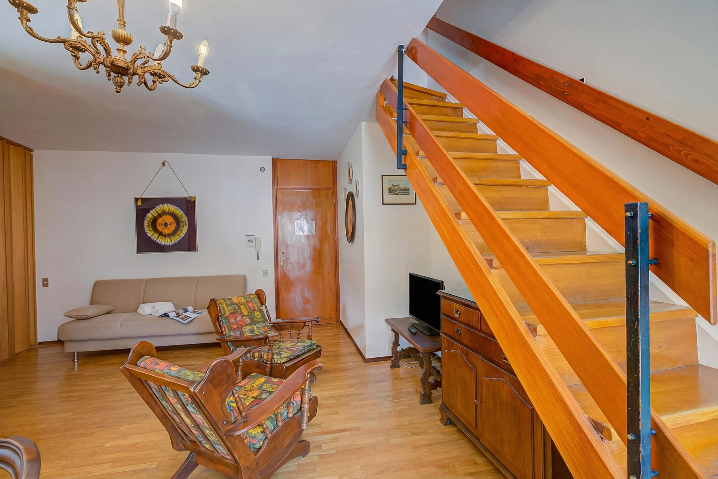 A living room with a wooden staircase, a sofa, two rocking chairs, a chandelier, and a television on a wooden cabinet.