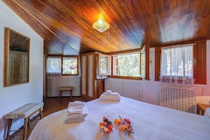 A room with a wooden ceiling, a bed with a white bedspread, a bench, a window with a view of trees, and a radiator.