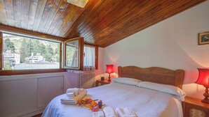 A bedroom with a wooden ceiling, a large bed with white linens, a window with a view of buildings, and a red lamp on the nightstand. - Il Rododendro  (City Center) (Bardonecchia)