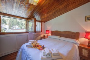Un dormitorio con techo de madera, una cama grande con ropa blanca, una ventana con vistas a edificios y una lámpara roja en la mesita de noche.