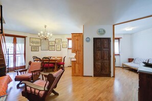 A spacious living room with a wooden floor, a dining area with a table and chairs, a chandelier, and a large wooden door.