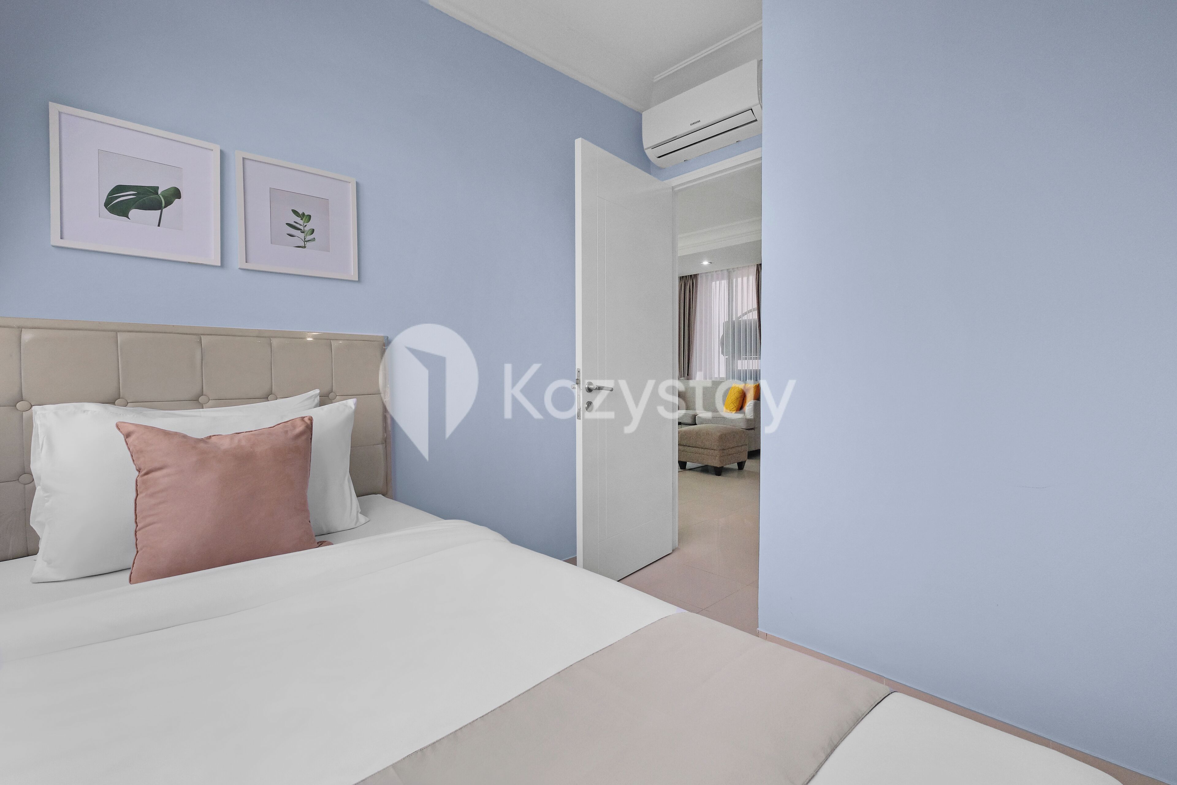 A hotel room with a bed, a headboard, a wall-mounted air conditioner, and two framed botanical prints.