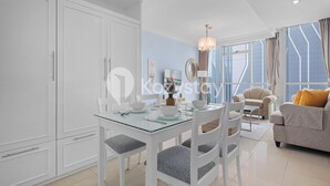 Un salon moderne avec une table à manger en verre, des meubles blancs, un canapé et de grandes fenêtres ornées de rideaux.