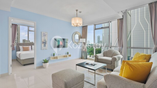 Un salon moderne avec un canapé, un fauteuil, un pouf et une table basse. On aperçoit un lit au fond et une grande fenêtre avec des rideaux.