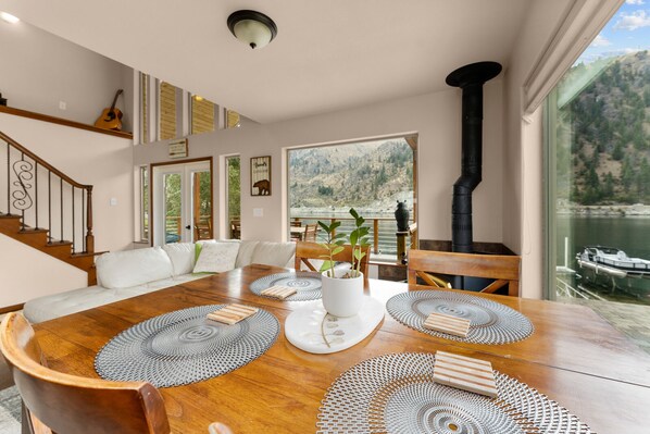 A spacious dining area with a wooden table, surrounded by chairs, and a large window offering a view of a lake and mountains. - Riverfront: Private Beach, Hot Tub, Sauna, Dock & Boat Launch! (Chelan)