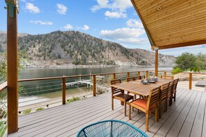 A wooden deck with a table and chairs overlooking a lake and mountains. - Riverfront: Private Beach, Hot Tub, Sauna, Dock & Boat Launch! (Chelan)