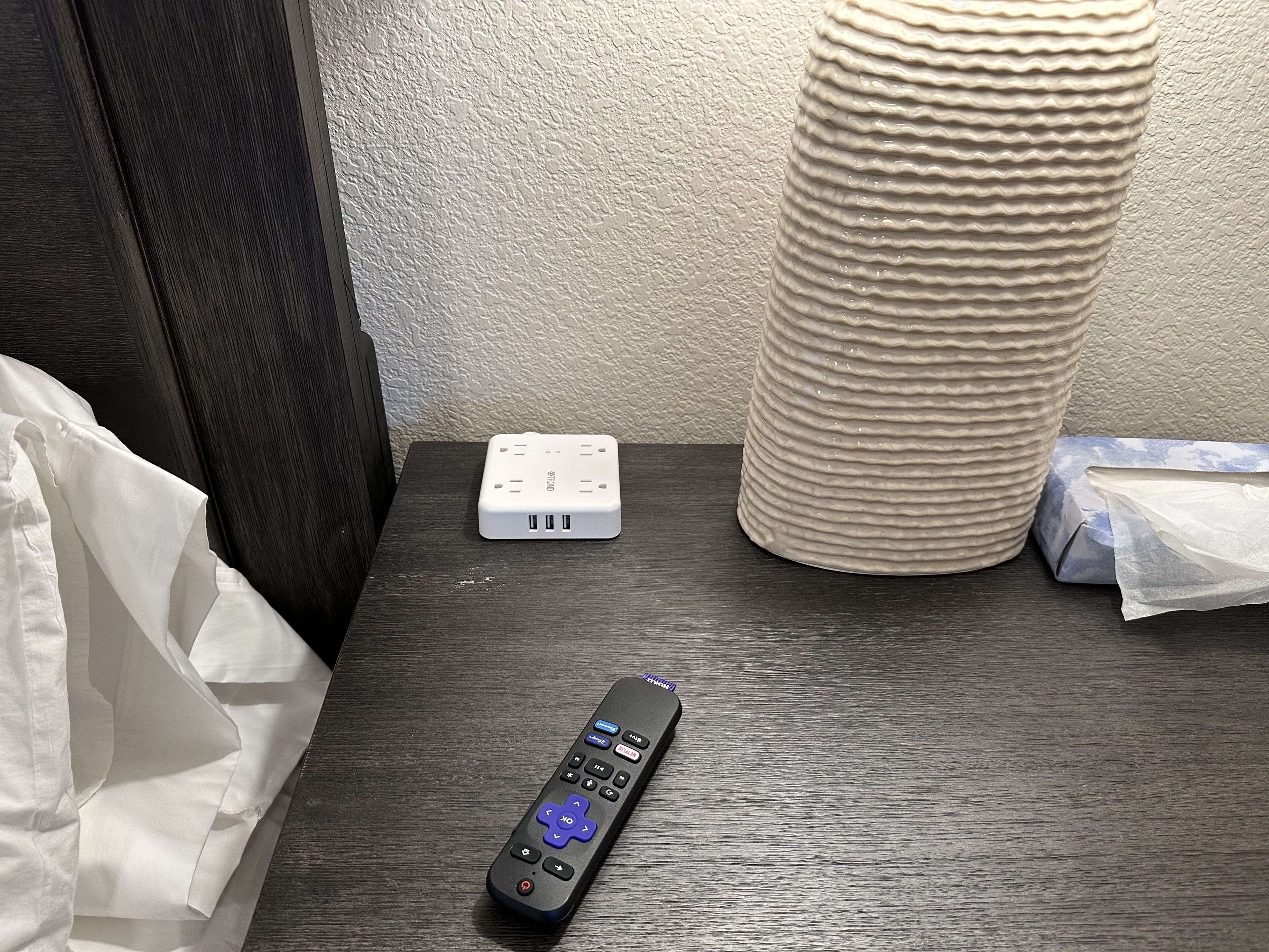 A remote control, a power strip, and a tissue box on a wooden nightstand.