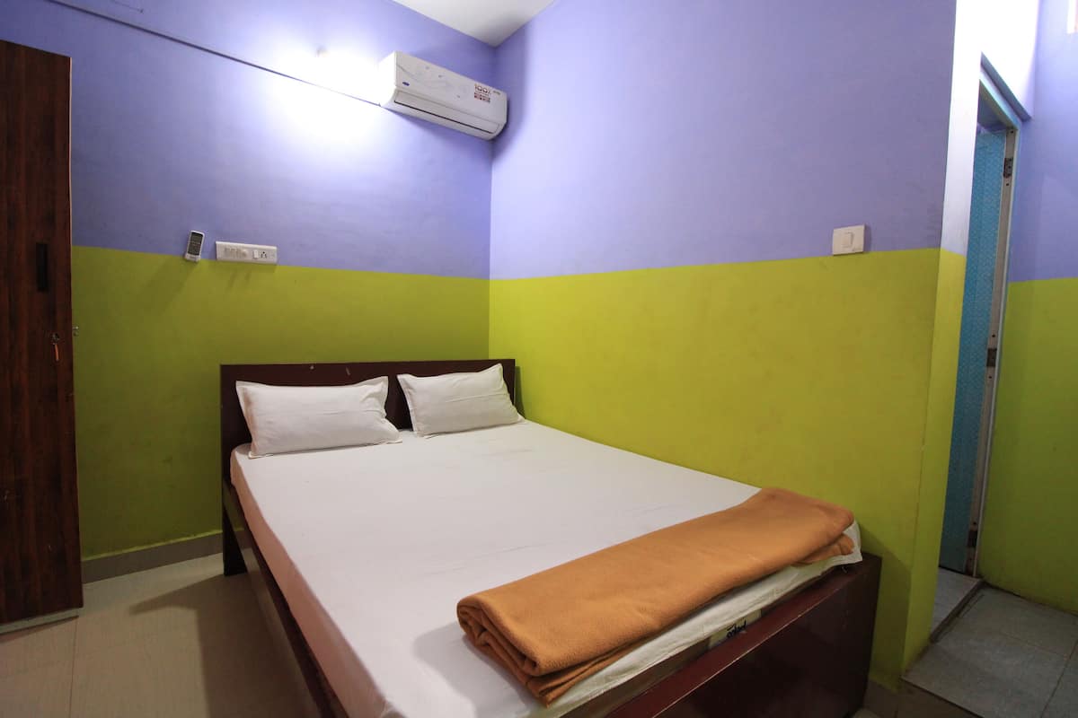 Uma cama de solteiro com colchão branco e duas almofadas, um cobertor marrom dobrado ao pé da cama, e um aparelho de ar condicionado fixado na parede.