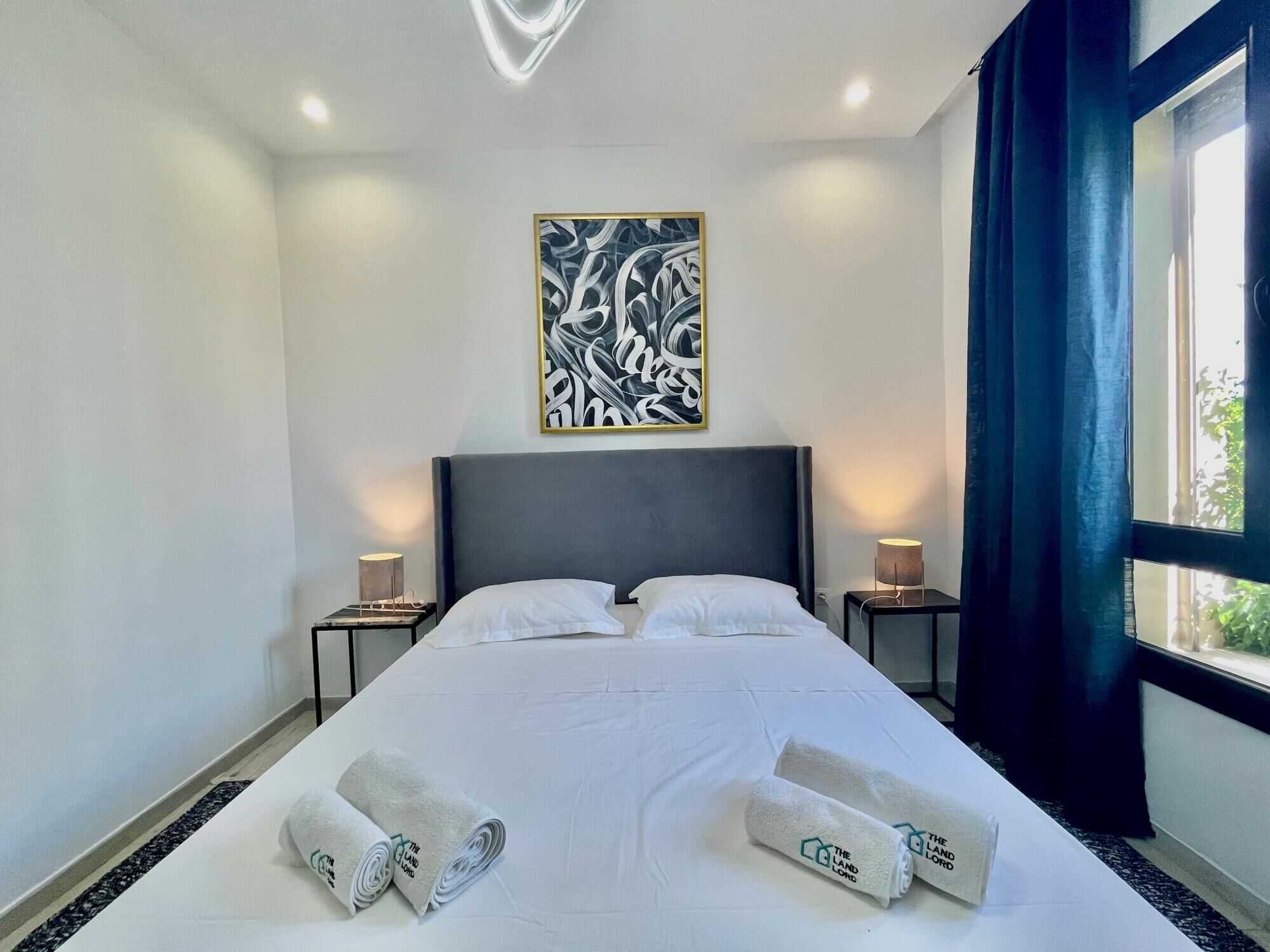 A hotel room with a large bed, two bedside tables with lamps, a wall-mounted abstract art piece, and a view through a window with curtains.