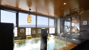 A hot spring bath area with a wooden floor, a central pillar, and a view of mountains through large windows.