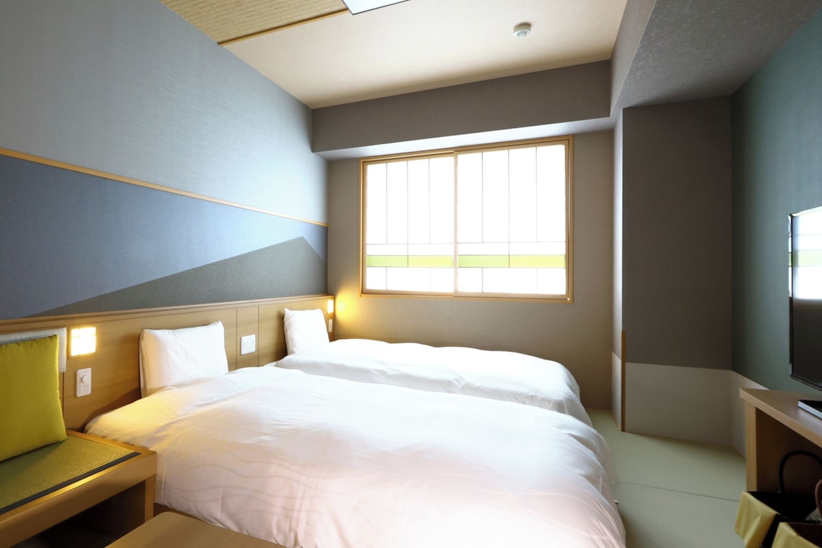 A hotel room with a large bed, a bedside table, a bench, and a window providing natural light.
