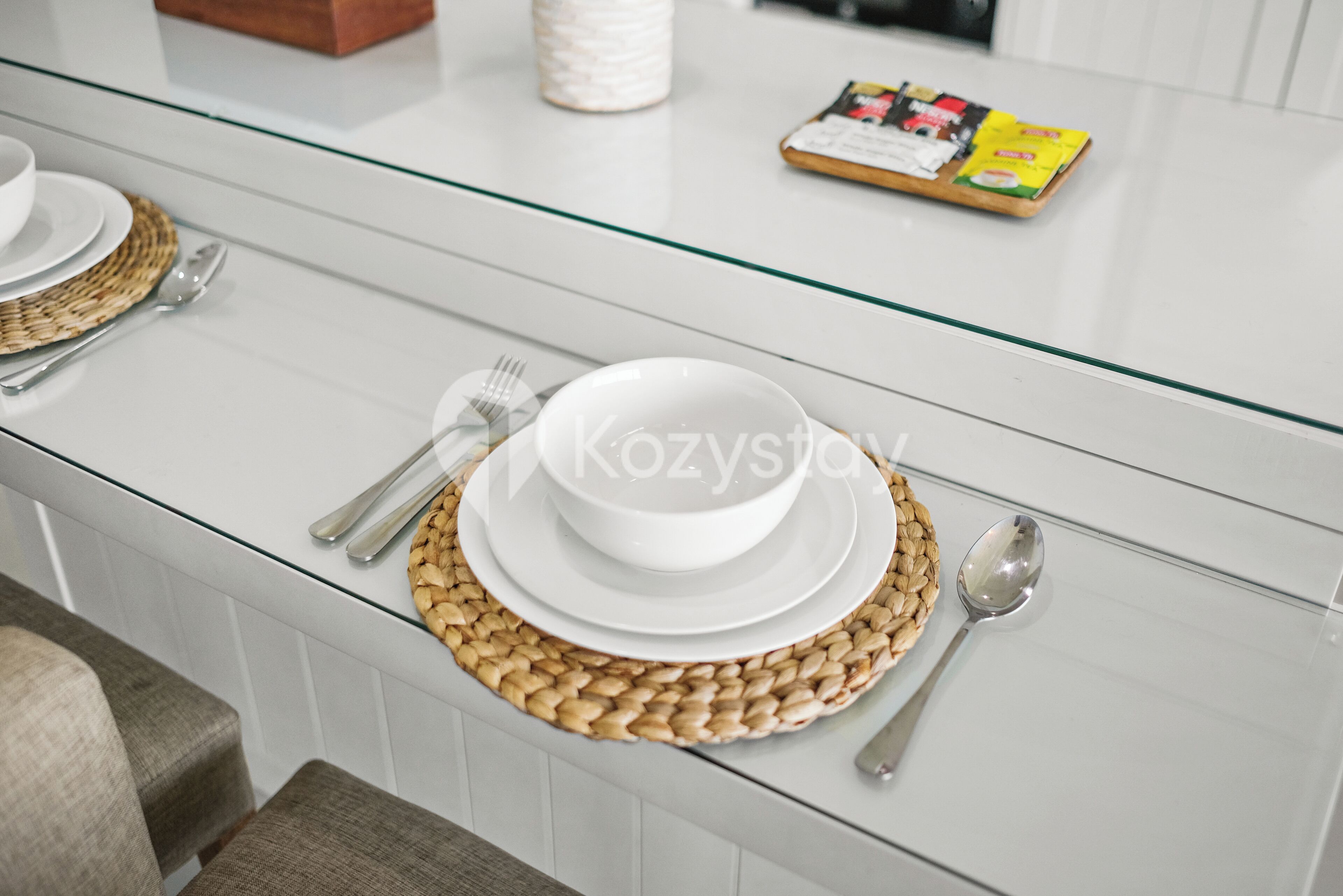 Un comedor con una mesa de vidrio, platos blancos, cubiertos y un posavasos tejido.