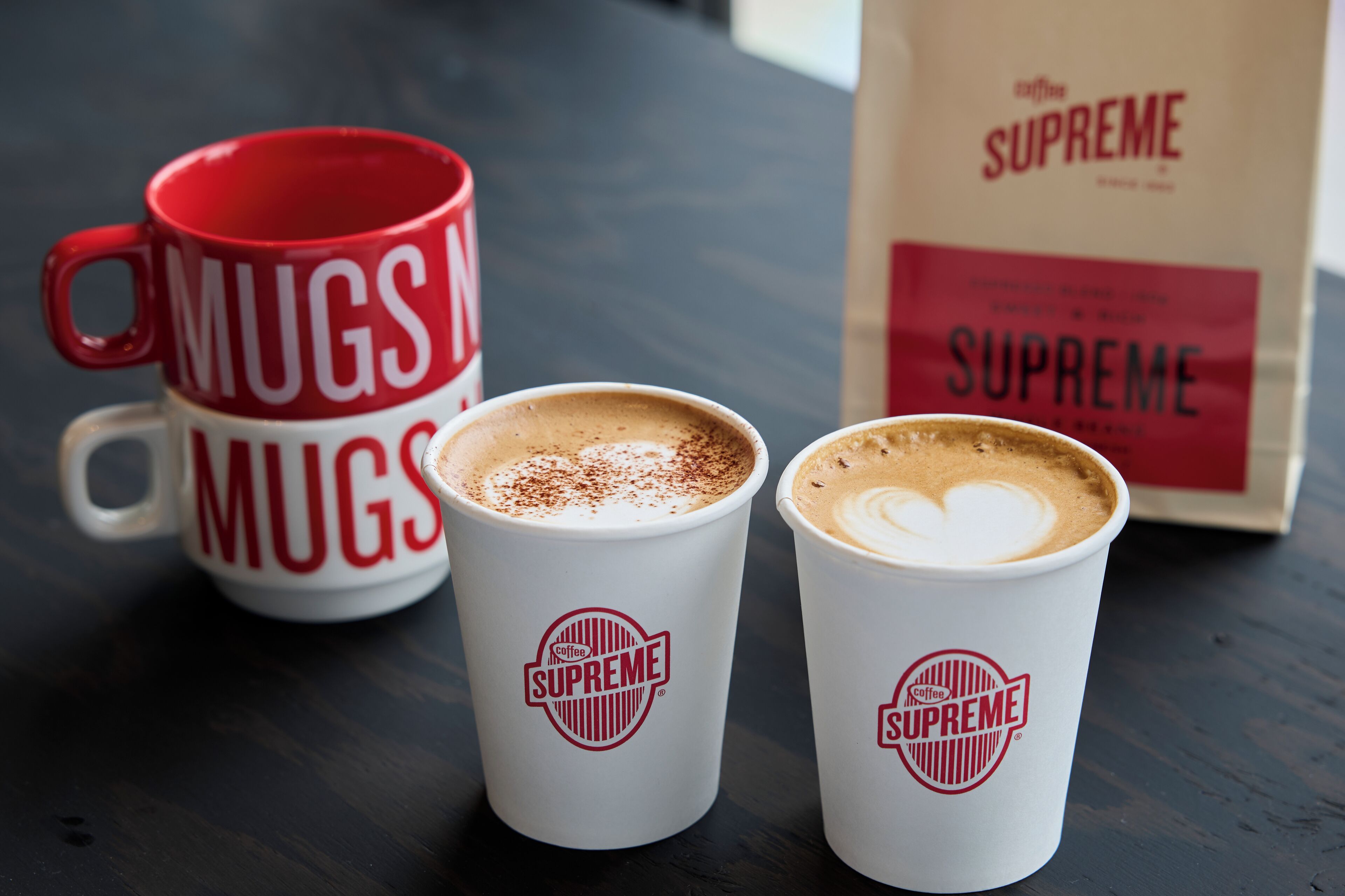 Dos tazas blancas de café con logotipos rojos y una taza roja con texto blanco.