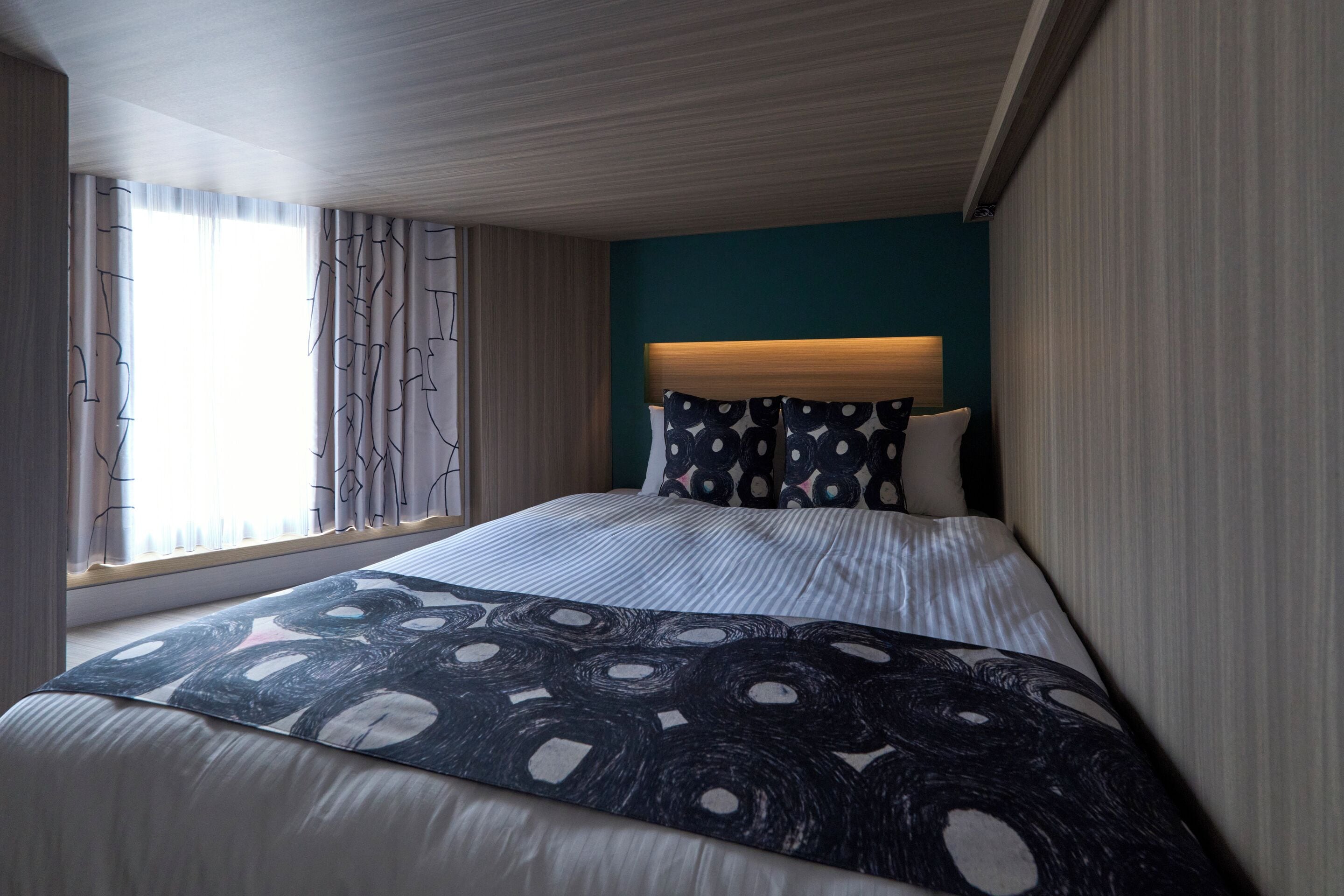 A modern hotel room with a large bed, a headboard with a light fixture, a window with curtains, and a teal accent wall.
