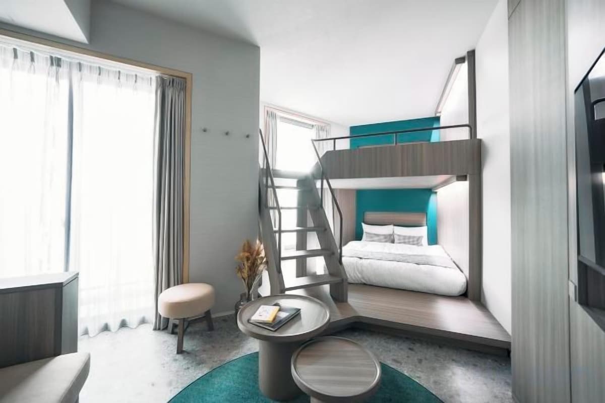 A compact hotel room with a bunk bed, a small seating area, and a window with curtains.