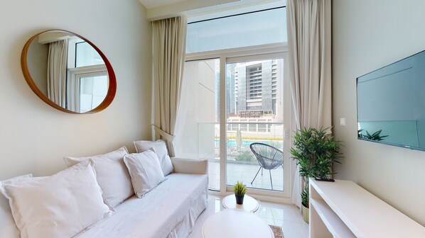 A modern hotel room with a white sofa, a round mirror, a flat-screen TV, and a balcony with a view of high-rise buildings.