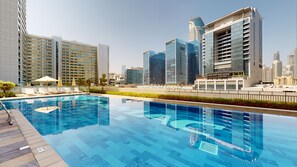 A modern hotel with a rooftop infinity pool, high-rise buildings, and a clear sky.