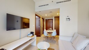 Una camera d'albergo moderna con una TV a schermo piatto fissata al muro, un divano bianco, un piccolo tavolino rotondo e un'area da pranzo con tavolo e sedie bianche.