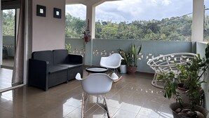 Un balcón con sofá de mimbre negro, sillas blancas y plantas en macetas.