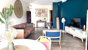 Una sala de estar con un sofá marrón, una silla azul, una mesa blanca y un televisor sobre un mueble blanco.