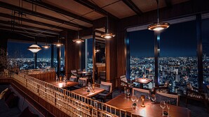 Un restaurante de altura con varias mesas y sillas, amplias ventanas que ofrecen vistas de la ciudad e iluminación colgante.