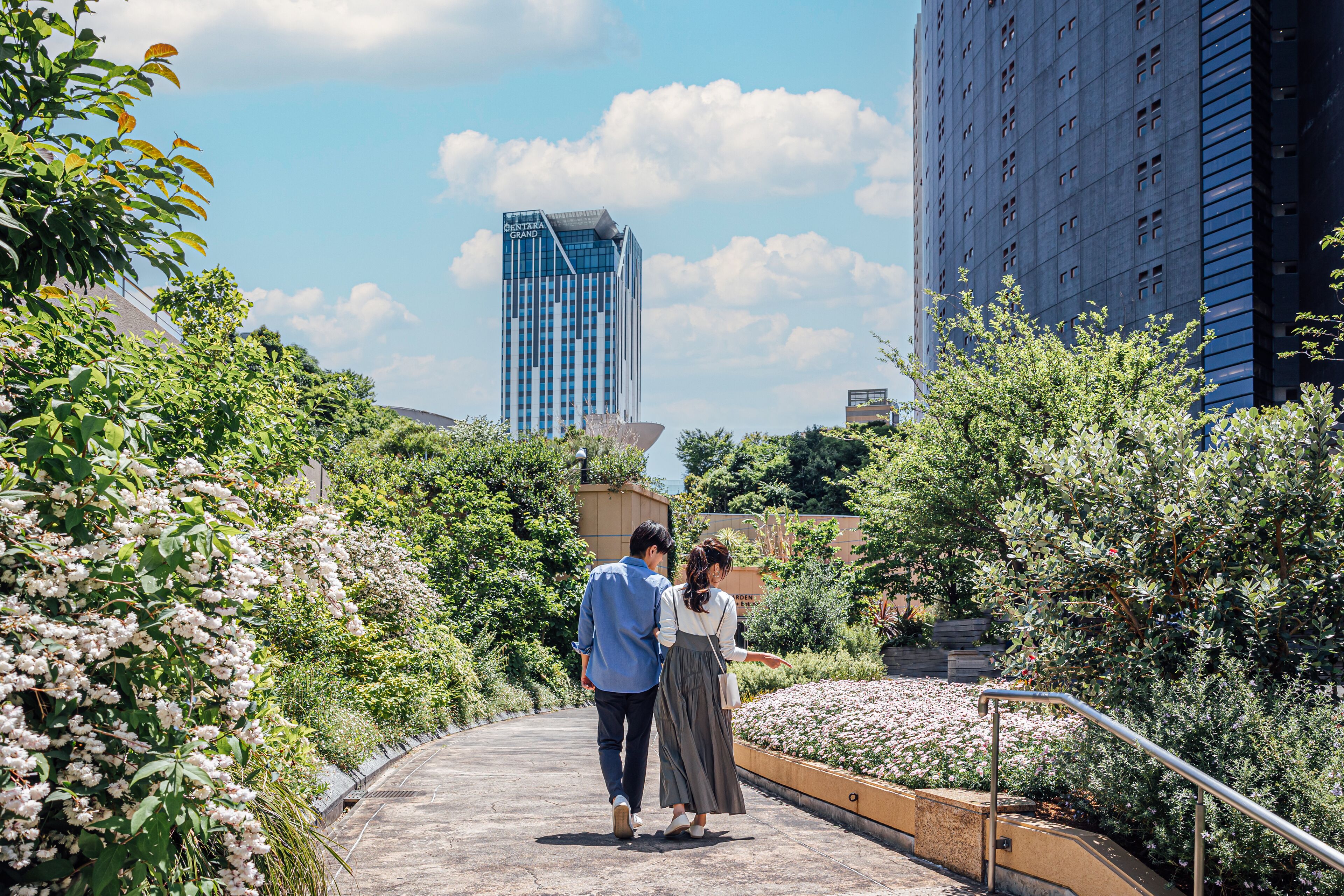 A couple walking on a pathway surrounded by greenery and buildings.