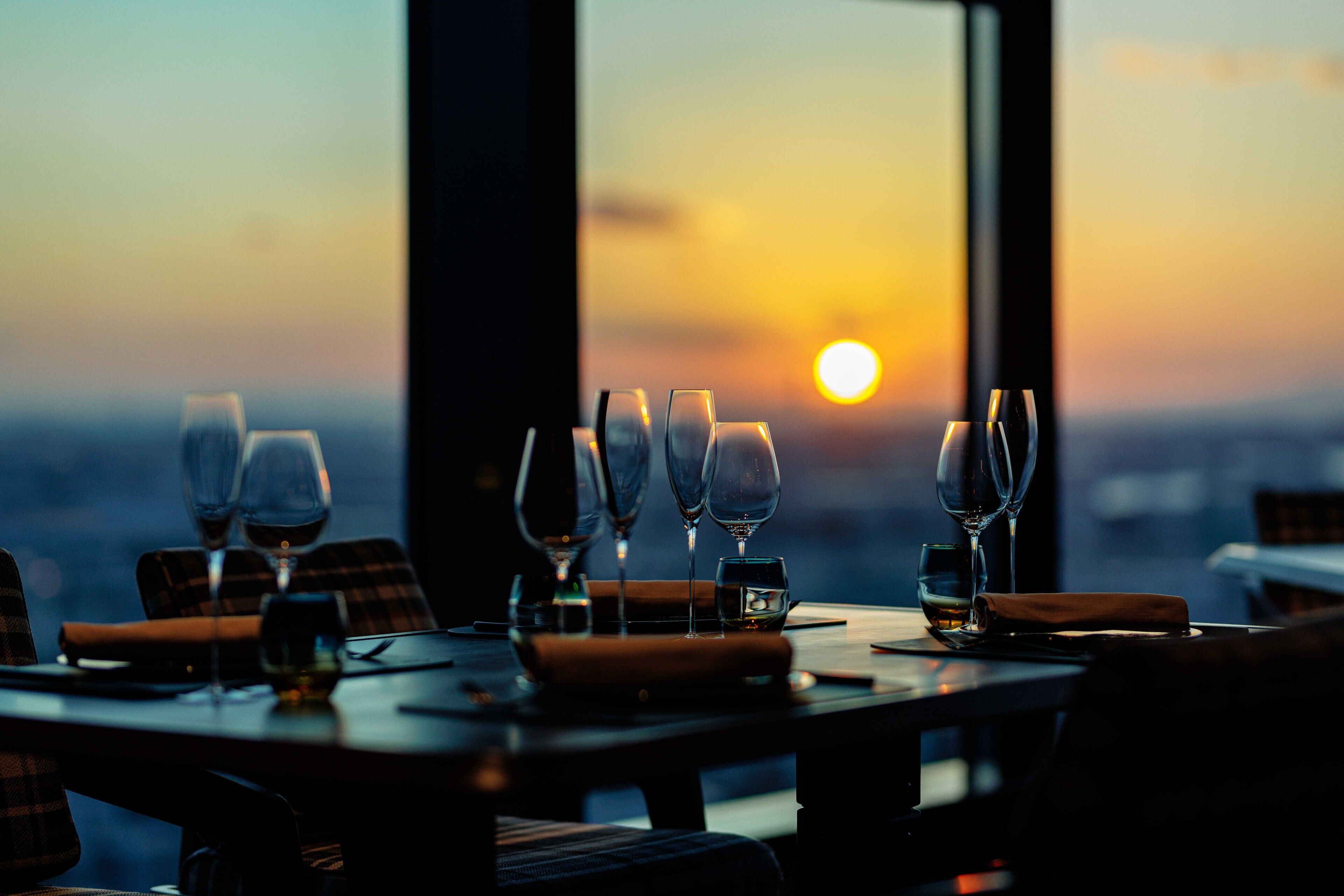 Una mesa de comedor con varias copas de vino y una servilleta, frente a una vista al atardecer a través de amplias ventanas.