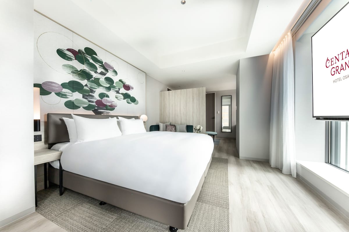 A modern hotel room with a large bed, a desk, a wall-mounted TV, and a view of the hotel name on the wall.