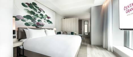 Habitación de hotel moderna con una cama grande, un escritorio, un televisor montado en la pared y vistas al nombre del hotel en la pared.