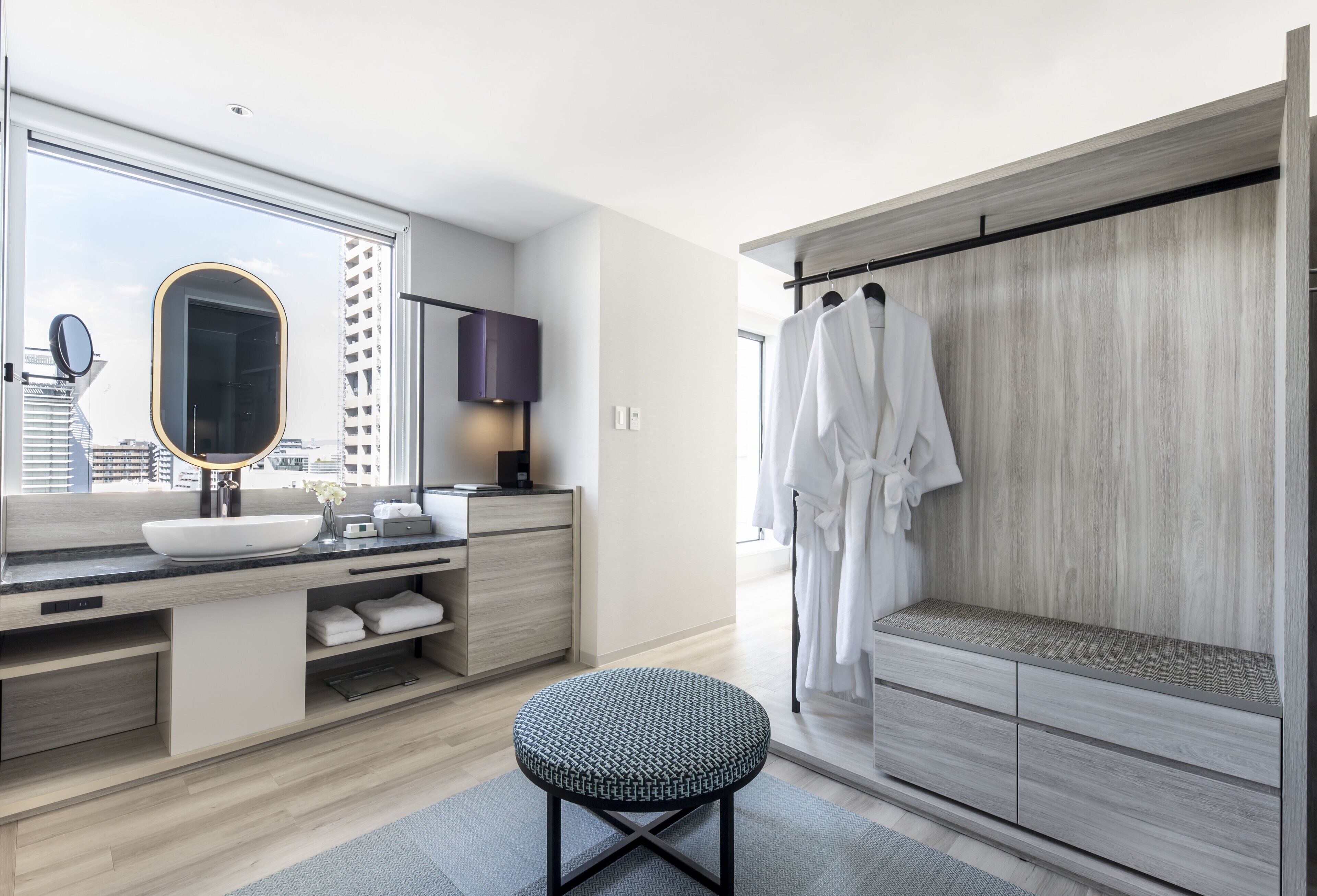 A modern bathroom with a large mirror, a round sink, a vanity with storage, a round stool, and a walk-in shower with a wooden panel wall.