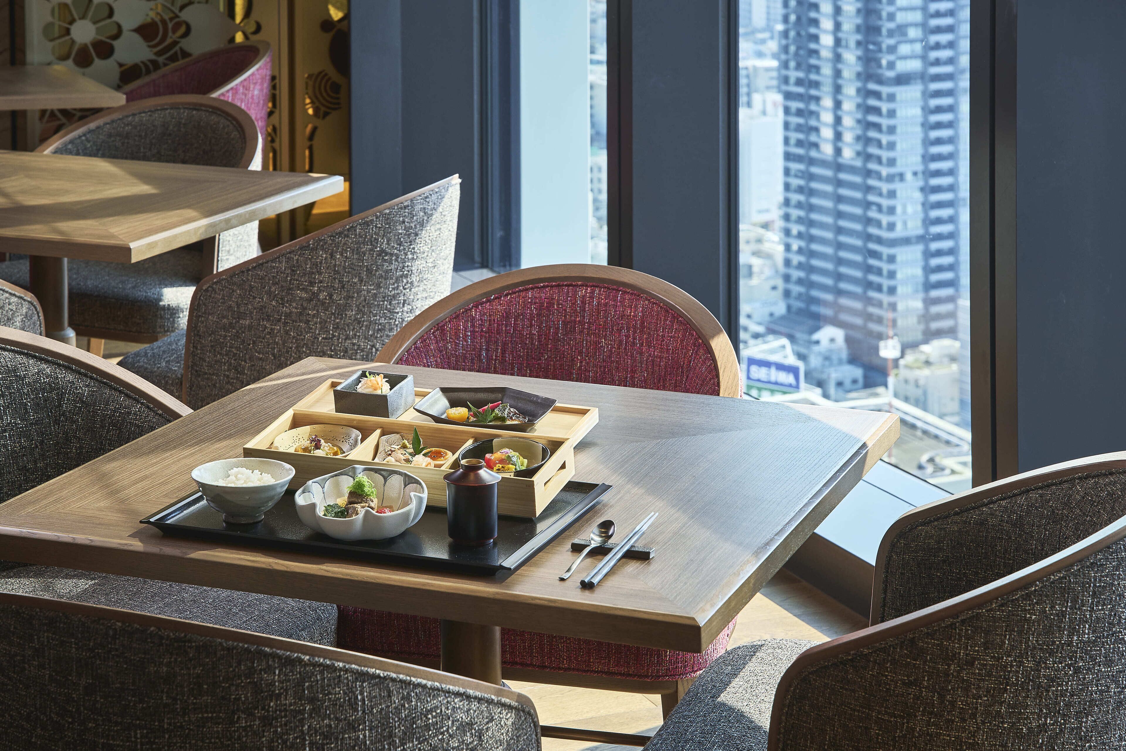 Un comedor con una mesa lista para una comida, que incluye tazones de arroz y platos, y vista a un paisaje urbano a través de amplios ventanales.