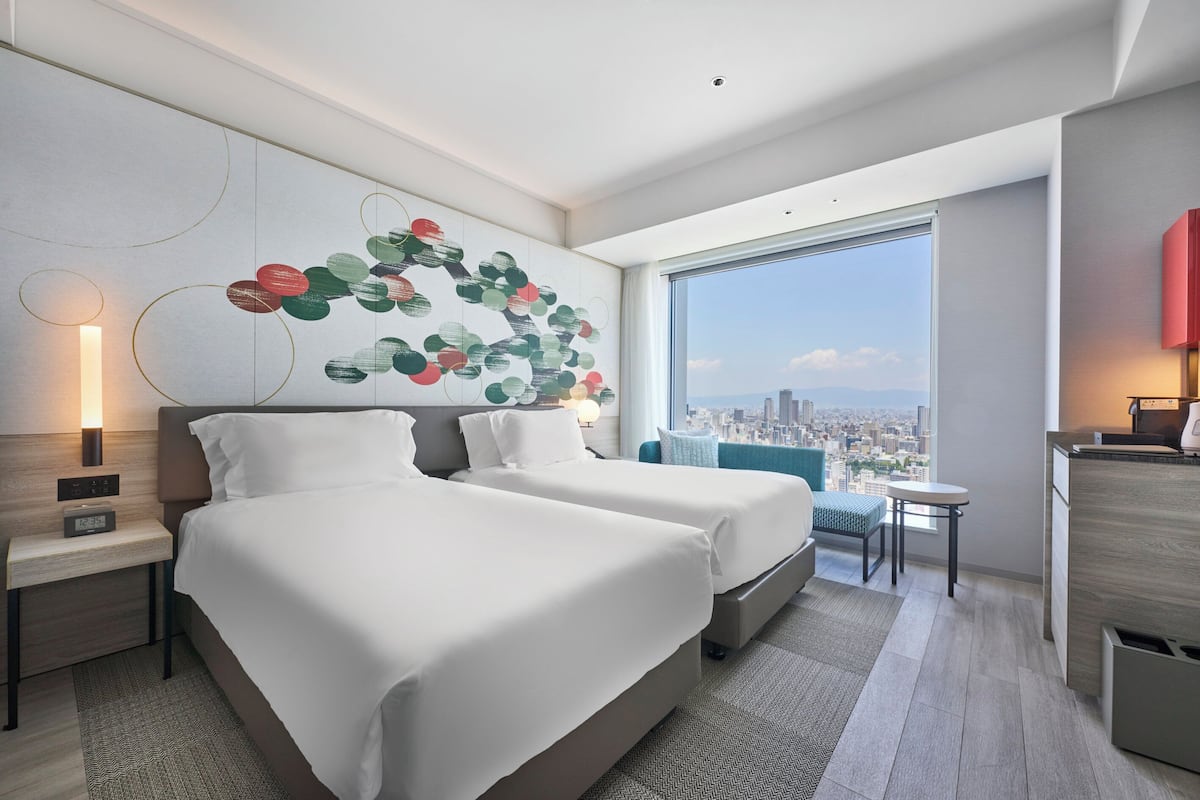 A modern hotel room with two beds, a desk, a chair, a small table, and a large window offering a city view.