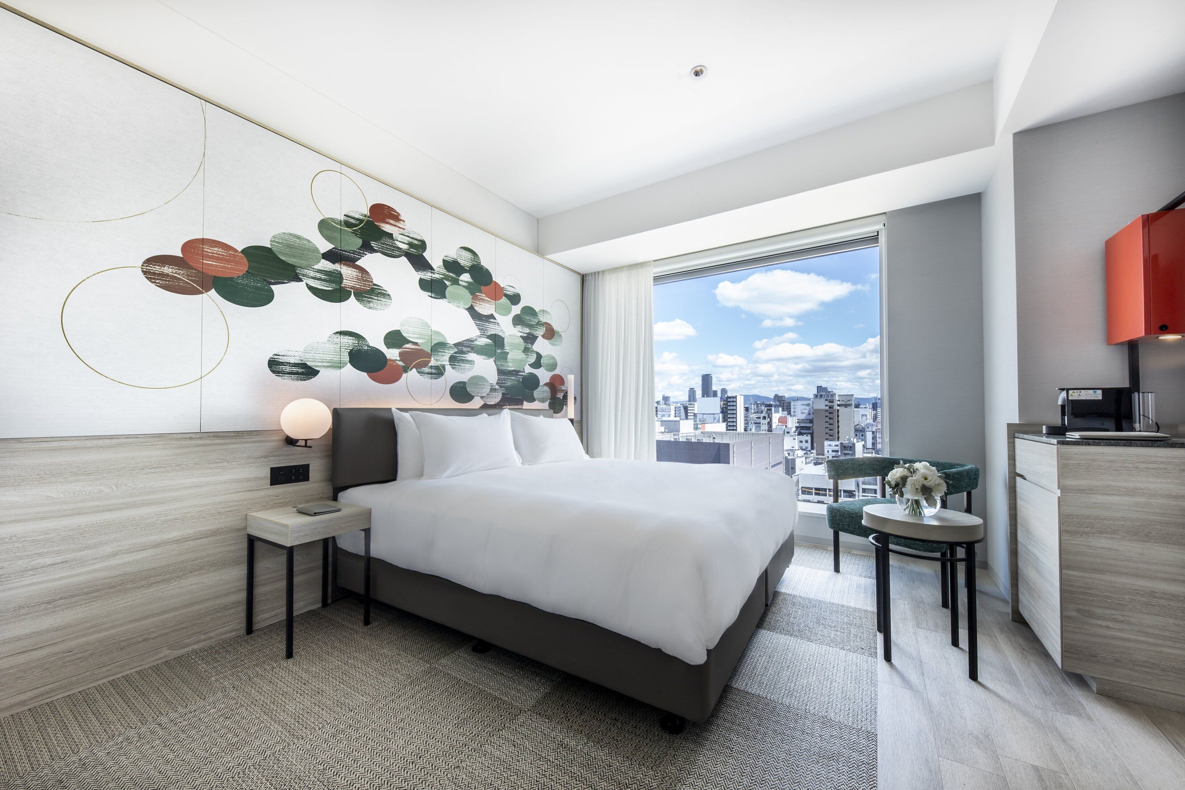 A modern hotel room with a large bed, a city view, a small dining area, and a decorative wall art piece.