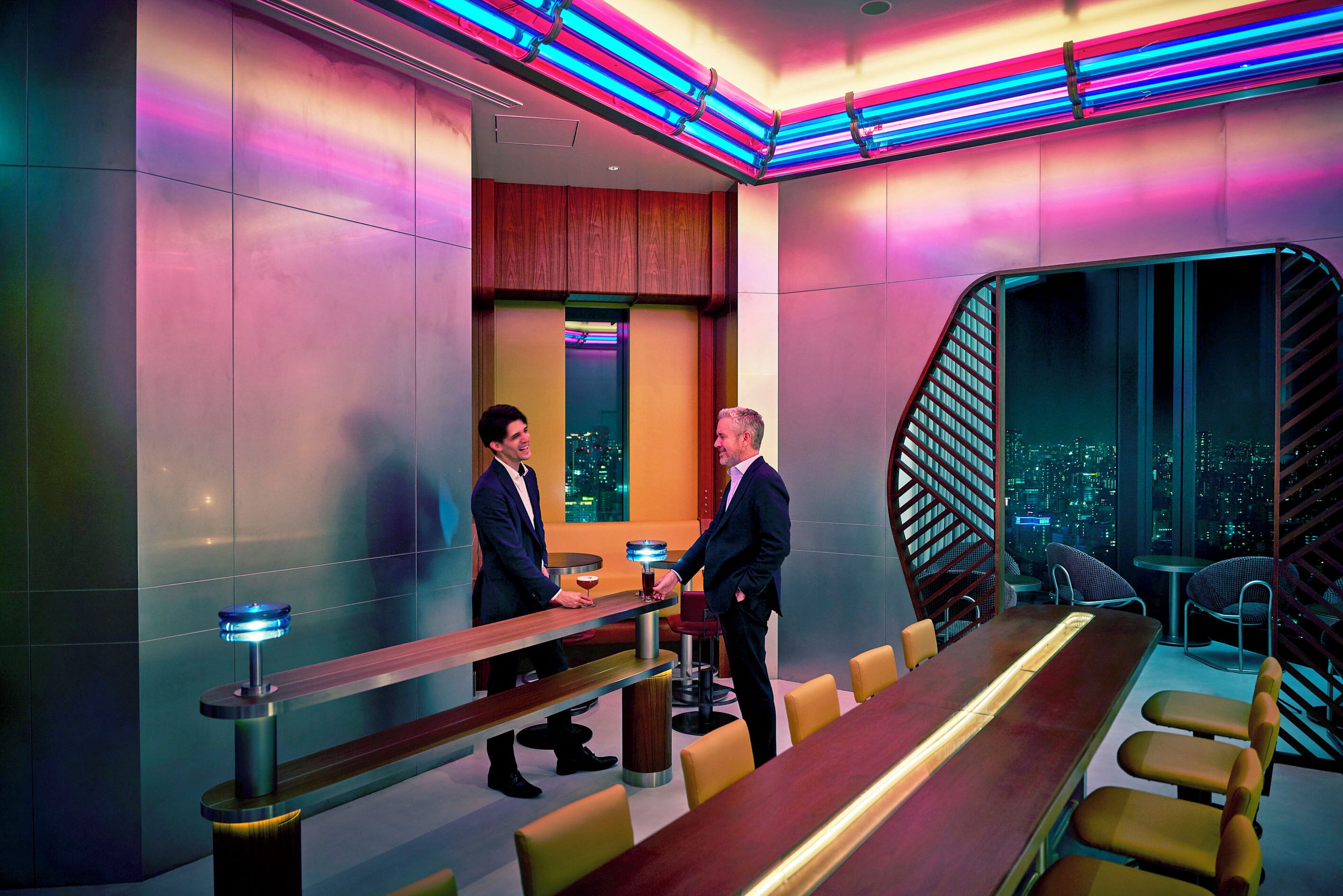 A modern hotel lobby with a long reception desk, seating area, and a view of the cityscape through large windows.