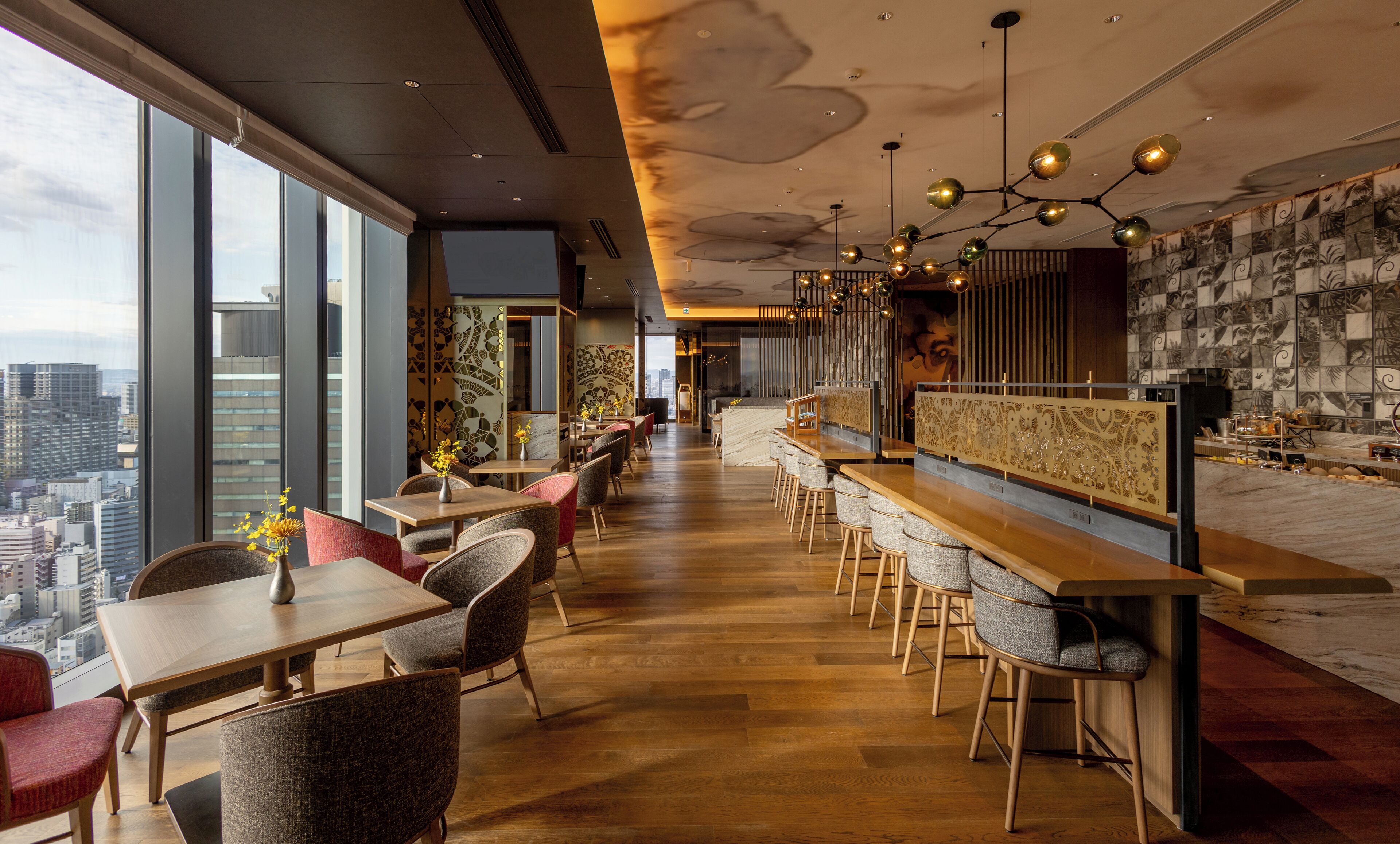 A modern bar area with wooden tables and chairs, a high ceiling with pendant lighting, and a city view through large windows.