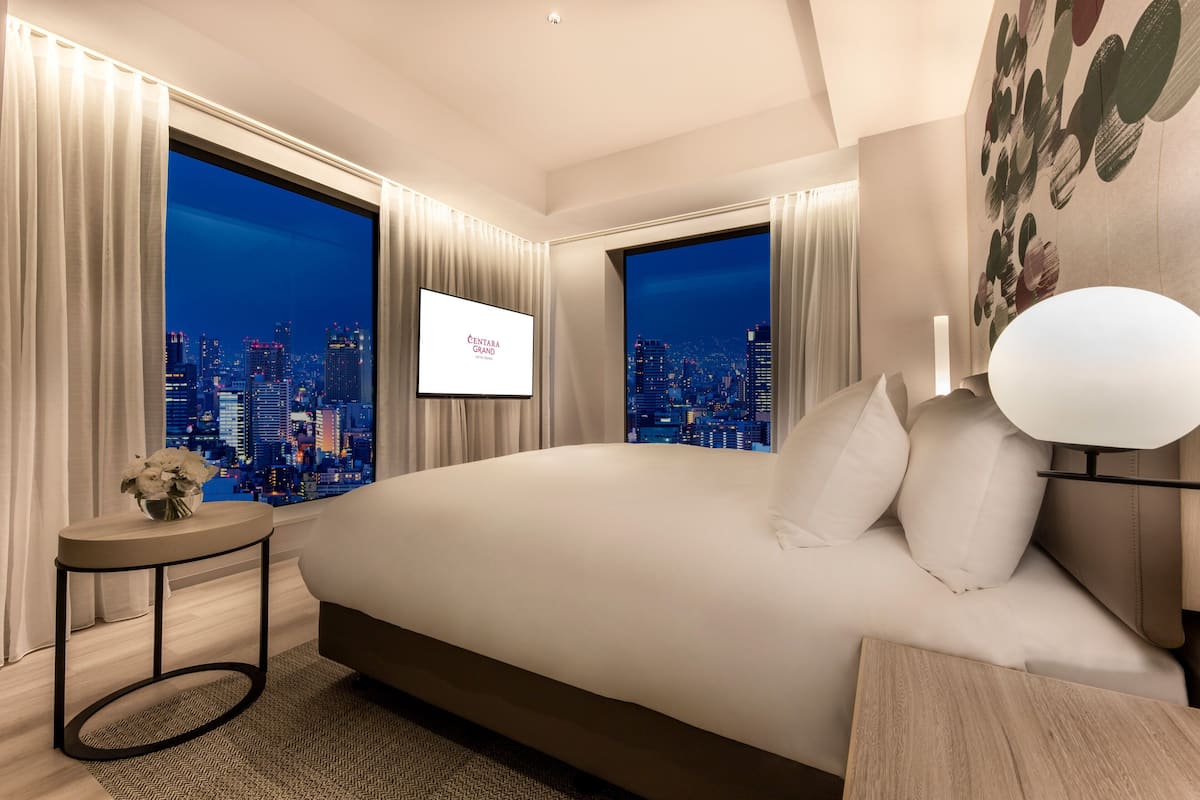 A modern hotel room with a large bed, a TV on the wall, and a cityscape view through the windows.
