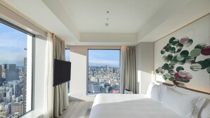 Una habitación de hotel moderna con una cama grande, un televisor de pantalla plana y vistas a la ciudad a través de amplias ventanas.