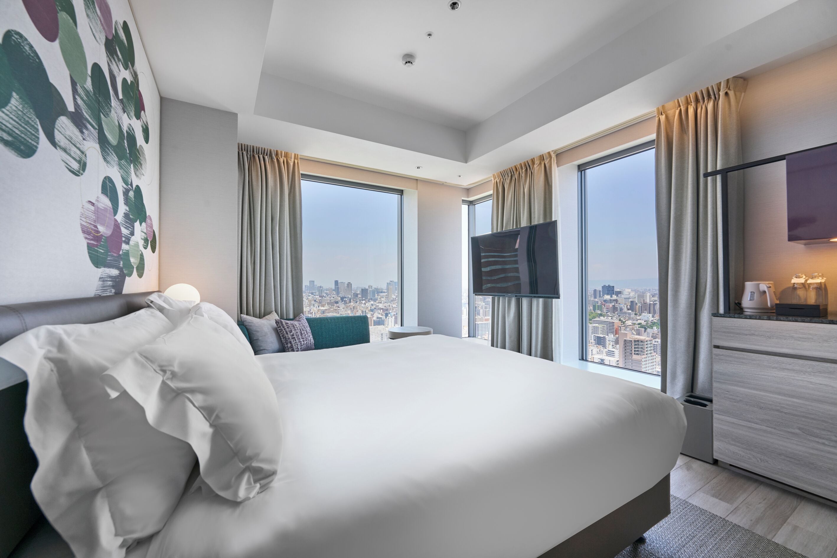 A modern hotel room with a large bed, a flat-screen TV, a city view, and a minimalist kitchenette.